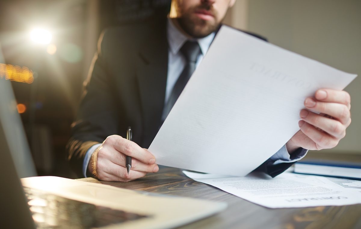 Portrait of modern bearded businessman holding papers in hands, reading and analyzing contract documentation at desk with laptop